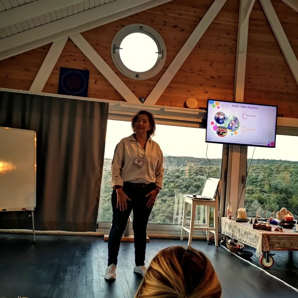 A teacher speaking in a wooden hall with a round window and forest views