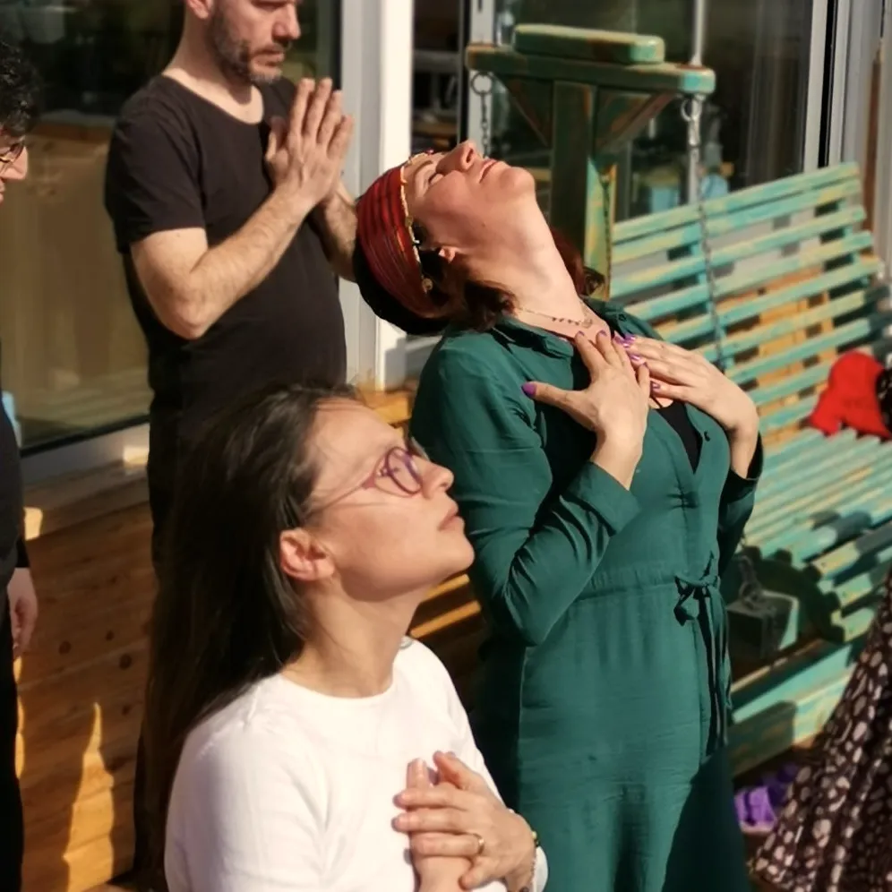Güneşli bir verandada gözleri kapalı, elleri göğsünde nefes alan iki kadın ve arkalarında avuçlarını birleştirmiş bir adam